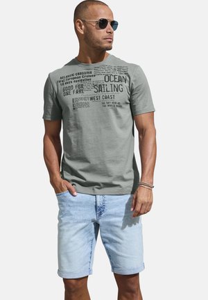Mann mit Sonnenbrille, grauem Grafik-T-Shirt mit Segel-Text, hellblauen hochgekrempelten Jeansshorts und Armbändern, steht mit einer Hand in der Tasche.