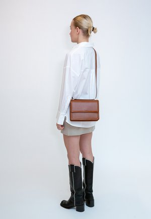 Donna con capelli biondi raccolti in un chignon, indossa una camicia bianca, una gonna beige, stivali neri al ginocchio e porta una borsa a tracolla in pelle marrone.