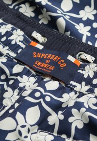 Bañador navy con patrón floral blanco, que cuenta con una cintura con cordón y una etiqueta de marca en naranja y azul. También incluye forro de malla.