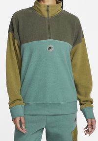 Fleece-pola med halvdragkedja; har gröna och teal-färgblock, runt logotypdetalj och ribbade ärmslut. Mjuk, texturerad tyg.