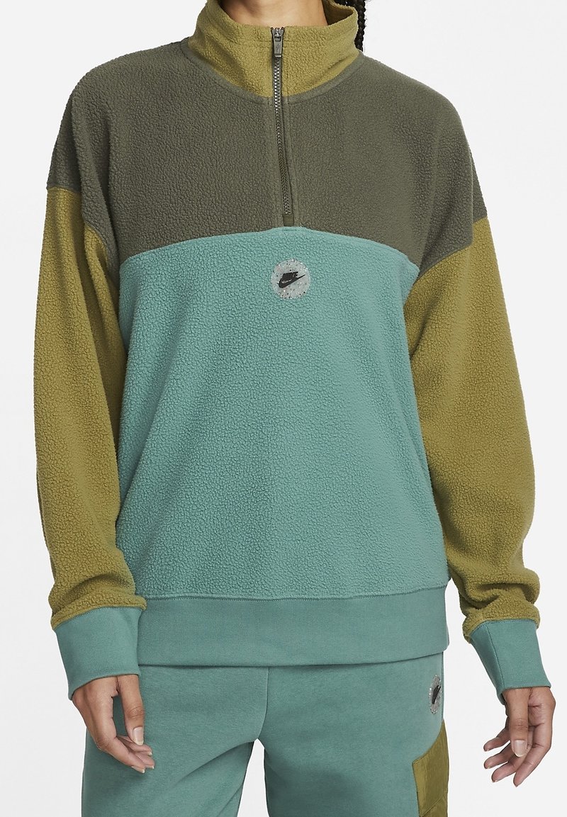 Fleece-pola med halvdragkedja; har gröna och teal-färgblock, runt logotypdetalj och ribbade ärmslut. Mjuk, texturerad tyg.