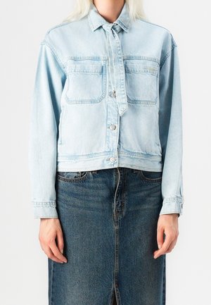 Person iført en lyseblå denimjakke med forlommer og knapper kombineret med et mørkeblå denimskørt med frontslids.