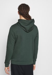 Tmavě zelená mikina s kapucí vyrobená z měkkého materiálu, s nastavitelnou kapucí a žebrovanými manžetami. Disponuje jednoduchým, čistým designem bez viditelných log.