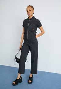 Salopette en denim noir à manches courtes, fermeture éclair à l'avant, deux poches poitrine, ceinture à la taille et coupe droite. Portée avec des chaussures à enfiler à plateforme.
