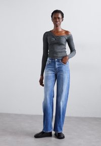 Woman standing against plain wall wearing grey off-shoulder long sleeve top, blue jeans, and black shoes with one hand in pocket.