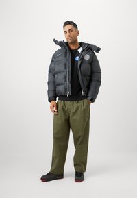 Chaqueta acolchada negra con un cuello alto y un logo, llevada sobre una camisa negra, combinada con pantalones de pierna ancha en verde oliva y zapatos negros.