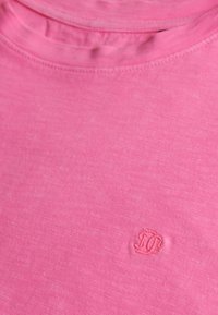 Close-up van een roze T-shirt met een ronde halslijn en een klein geborduurd logo op de borst.