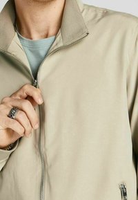 Bunda na zip v světle béžové látce, s límcem na stojáka, boční kapsou na zip a hladkou texturou. Ruka osoby je blízko zipu.