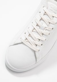 White leather sneaker with a smooth texture, round toe, and flat laces, featuring subtle stitching details and a rubber outsole.