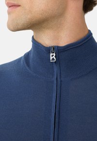 Mann in einem blauen Strickpullover mit Reißverschluss und hohem Kragen, mit einem Reißverschlusszieher in Form des Buchstabens "B."