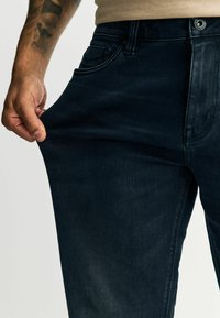 Tumed sinised teksapüksid kitsas lõikega, nähtavad taskudetailid ja käsi, mis hoiab kanga venitamist ja tekstuuri näitamiseks.