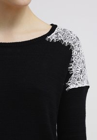 Pull en tricot noir avec une superposition de dentelle florale blanche sur l'épaule et le haut du bras, porté par une personne à la peau claire.