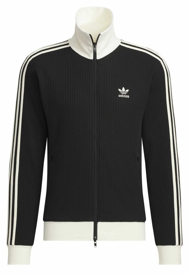 Chaqueta negra de cremallera con textura acanalada, acentos blancos, cuello alto y bolsillos laterales. Presenta un diseño de tres franjas blancas en las mangas y el logo de Adidas.