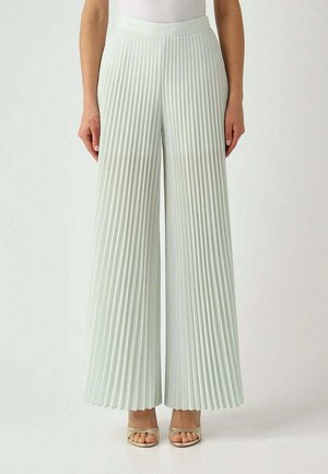 Donna indossa pantaloni plissettati a vita alta e gamba larga di colore pastello chiaro con tacchi open-toe, in piedi su uno sfondo semplice.