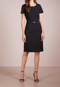 Robe noire mi-longue avec manches courtes, col rond et taille cintrée, ornée d'un accent argenté à la taille. Texture lisse.