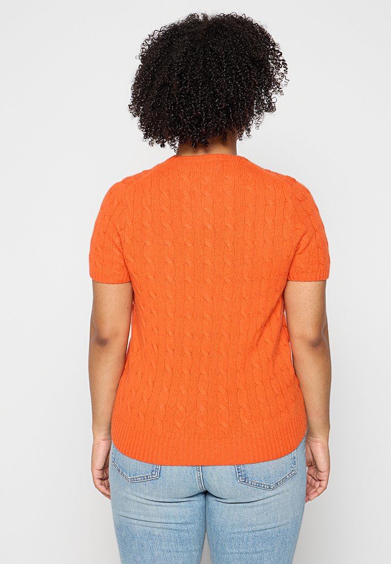 Maglione corto a maniche corte di colore arancione, caratterizzato da un motivo a treccia e un colletto rotondo, abbinato a jeans in denim blu.