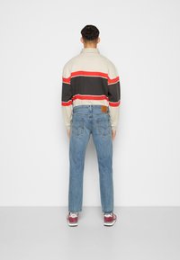 Hombre de pie frente a una pared blanca, vistiendo una camiseta de manga larga a rayas en tonos beige, negro y rojo, jeans azules y zapatillas color burdeos y blancas.