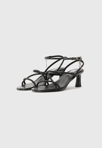 Sandales noires à talons hauts avec des fines lanières, dotées d'une texture brillante et d'une bride de cheville réglable avec un détail en chaîne décoratif.