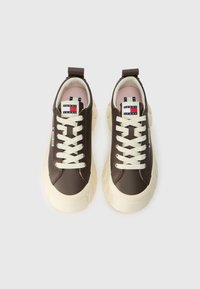 Smeđi tenisice s glatkom gornjom površinom, kremastim vezicama i debelim, teksturiranim kremastim potplatima. Imaju oznaku s logotipom na jeziku i petlju za jednostavno oblačenje.