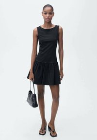 Black sleeveless dress with a fitted bodice and flared skirt. Features smooth fabric, knee-length hem, and worn with black flip-flops and a handbag.