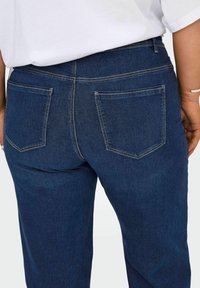 Tumši zili džinsi ar vidēju vidukli, divām aizmugurējām kabatām un kontrastējošu šuvju šuvēm. Gluda tekstūra un klasiskā piegriezuma.