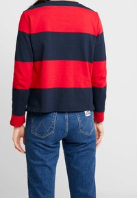 Maglietta a righe rossa e blu a maniche lunghe con collo rotondo. Tessuto morbido, vestibilità comoda, abbinata a jeans blu con dettaglio tasca sul retro.