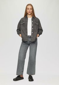 Schwarze Jeansjacke mit Knopfverschluss und zwei Brusttaschen, kombiniert mit grauen weit geschnittenen Jeans und schwarzen Slipschuhen. Weißes T-Shirt darunter.