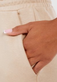 Joggers de tela beige con un bolsillo lateral, mostrando una mano descansando dentro. La textura parece suave con una cintura ligeramente elástica.