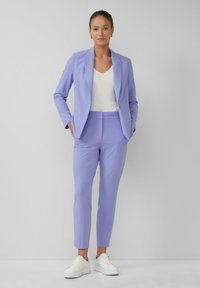 Helllila maßgeschneiderter Blazer und passende Hose aus glattem Stoff, kombiniert mit einem weißen Oberteil und weißen Sneakern, vor einem neutralen Hintergrund.