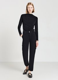 Schwarzes Langarmoberteil mit drapiertem Detail, kombiniert mit hochgeschnittenen schwarzen Cargo-Hosen mit Seitentaschen. Schwarze gemusterte Slip-On-Schuhe.