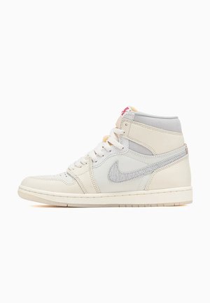 Zapatilla de corte alto en cuero color blanco roto y gris claro con un logo Nike gris texturizado y cordones blancos sobre un fondo blanco liso.