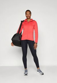 Rdeča jopa z zadrgo, črne pajkice, sive superge in črna športna torba, prikazane na modelu na nevtralnem ozadju.