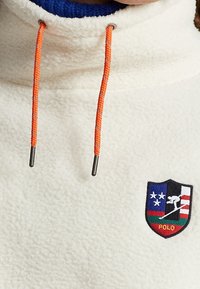 Fleece-pullover i krämfärg med hög krage, med oranga dragsnören och en lapp med flerfärgad design och texten "POLO".