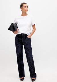 Musta corduroy-housut, joissa on sileä pinta ja leveät lahkeet, yhdistettynä valkoiseen puuvillateeseen ja mustaan clutch-laukkuun. Malli seisoo.