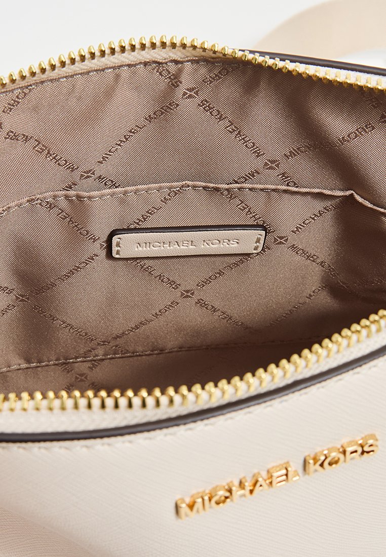 Interior de un bolso beige con tela clara que muestra un patrón de logotipos "MICHAEL KORS". Presenta un cierre con cremallera y una etiqueta de marca metálica.