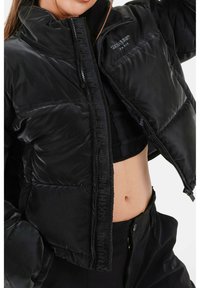 Zwarte pufferjack met hoge kraag, ritssluiting en textuurstof. Bevat een merknaam langs de rits en heeft een cropped ontwerp.