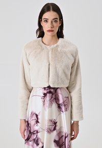 Veste en fausse fourrure cintrée dans un beige doux, avec un col rond, des manches longues et une texture subtile. Associée à une jupe à motif floral.