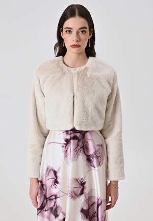 Kürzerer Kunstpelz-Jacke in sanftem Beige, mit rundem Hals, langen Ärmeln und subtiler Textur. Kombiniert mit einem Rock im Blumenmuster.