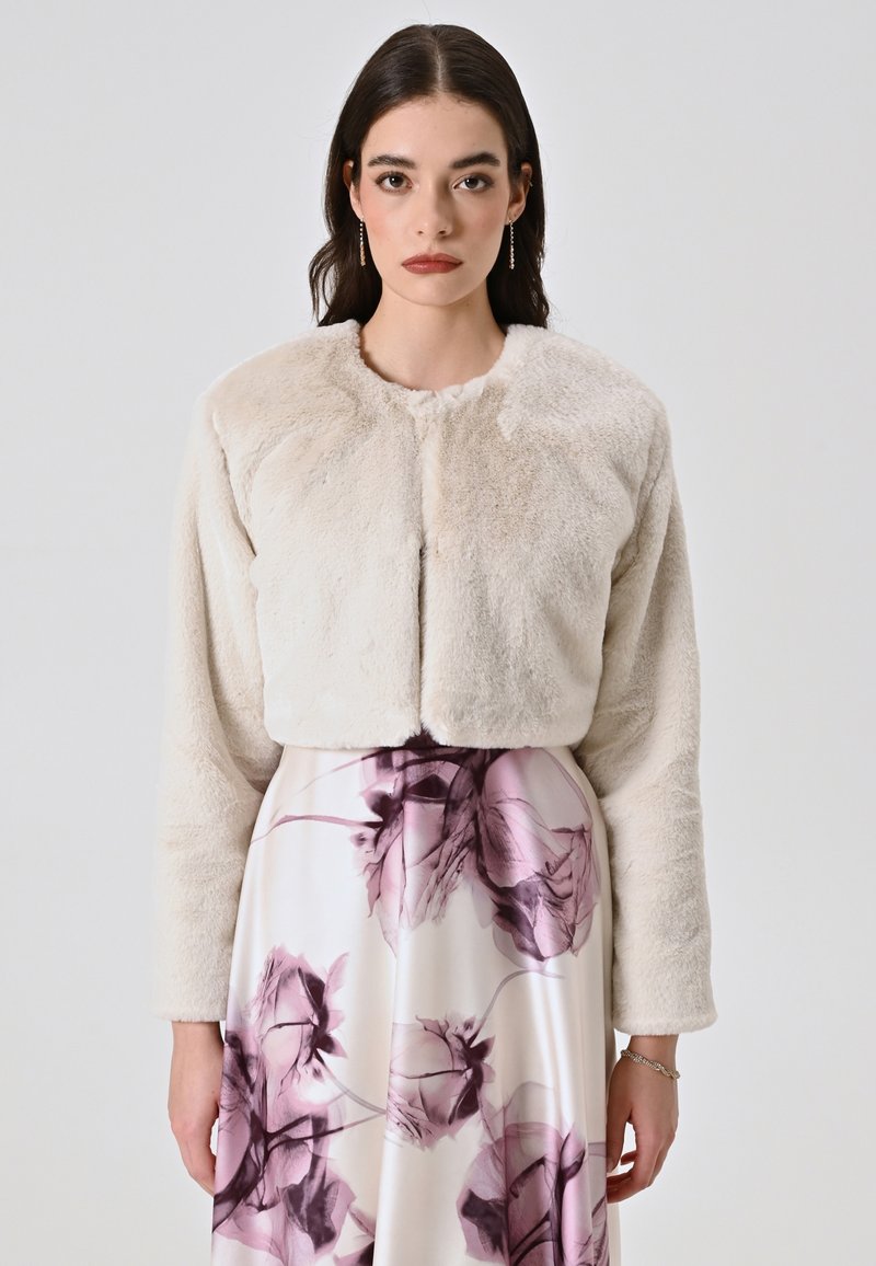 Veste en fausse fourrure cintrée dans un beige doux, avec un col rond, des manches longues et une texture subtile. Associée à une jupe à motif floral.