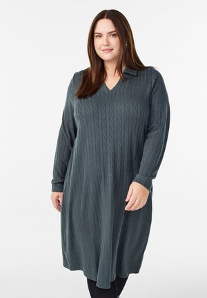 Langsleeve, v-hals jurk in donkergroen met een gestructureerd kabelbreipatroon. Heeft een ontspannen pasvorm en valt onder de knie.