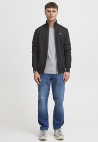 Schwarze Zip-up-Jacke mit hohem Kragen, graues T-Shirt darunter, blaue Jeans und hellfarbene Sneakers. Einfaches Design mit kleinem Logo.