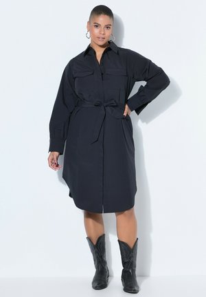 Kvinde i knælængde sort kjoleskjorte med bælte, lange ærmer, lommer og sorte cowboystøvler, stående med den ene hånd på hoften.