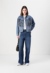 Jeansjacke mit Fransenverzierungen an der Vorderseite, kombiniert mit weiten Jeans. Das Model hält eine kleine olivgrüne Tasche. Weißer Hintergrund.