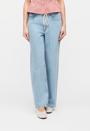 Persona vistiendo jeans anchos de mezclilla azul claro con cordón blanco y zapatos beige planos, combinados con una blusa a cuadros roja y blanca.