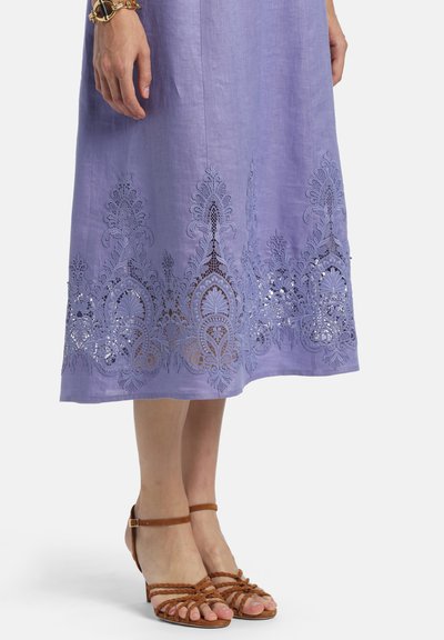 Lavendelfarbener Midirock mit aufwändiger Spitzenstickerei am Saum, kombiniert mit braunen, niedrigabsätzigen Riemchensandalen und einem goldenen Armband am linken Handgelenk.