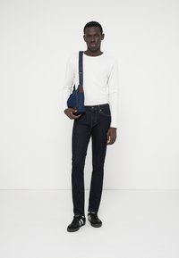 Calças de ganga escuras combinadas com uma camisa de manga longa branca e uma mala azul ao ombro. Ténis pretos com riscas brancas completam o visual.