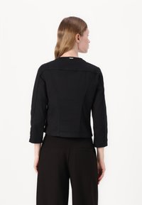 Giacca nera corta in cotone con maniche 3/4, dotata di colletto rotondo, cuciture a vista e dettagli hardware minimali sulla schiena.