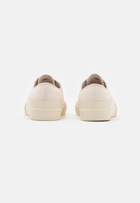 Beige plátěné tenisky s texturovaným povrchem, vybavené světle zbarvenou gumovou podrážkou, minimalistickým designem a bez viditelného branding na zadní straně.