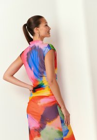 Frau mit braunen Haaren im Pferdeschwanz, die ein farbenfrohes, batikmusterndes Kleid trägt, steht mit einer Hand auf der Hüfte, lächelt und blickt an eine weiße Wand.