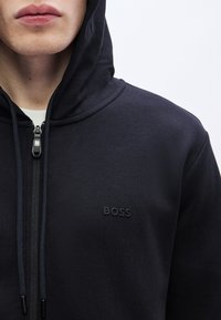 Črna majica s kapuco na zadrgo iz mehke tkanine, z zgornjo zadrgo, vrvico za zategovanje in majhnim vtisnjenim logotipom na levi strani prsnega koša.